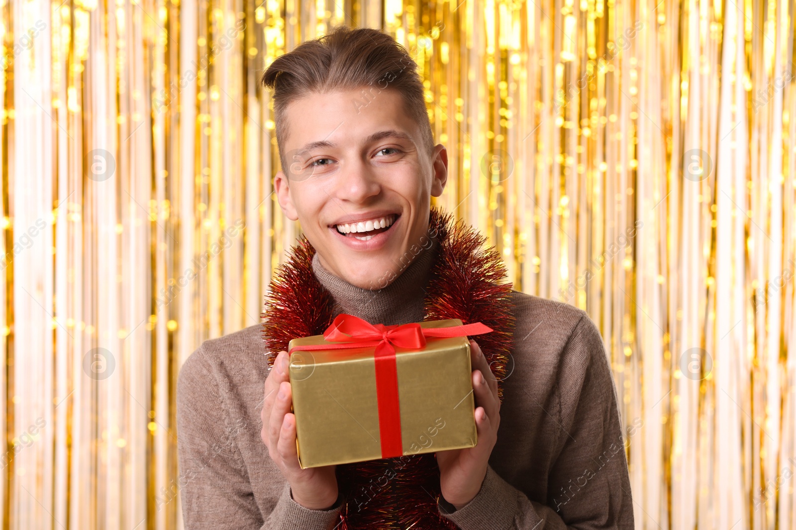 Photo of Happy young man with bright tinsel and gift box against foil curtain