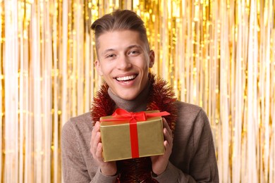 Photo of Happy young man with bright tinsel and gift box against foil curtain