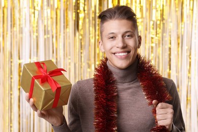 Photo of Happy young man with bright tinsel and gift box against foil curtain