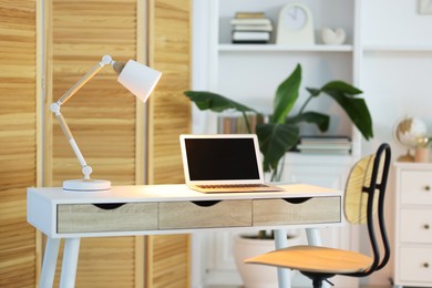 Comfortable workplace with laptop and lamp on table indoors