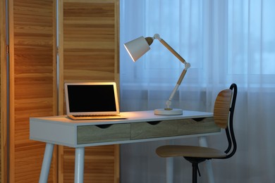 Photo of Comfortable workplace with laptop and lamp on table indoors