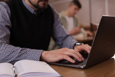 Photo of Man typing on laptop near book at desk in library, closeup