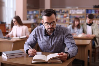 Photo of Handsome man reading book and taking notes at desk in public library