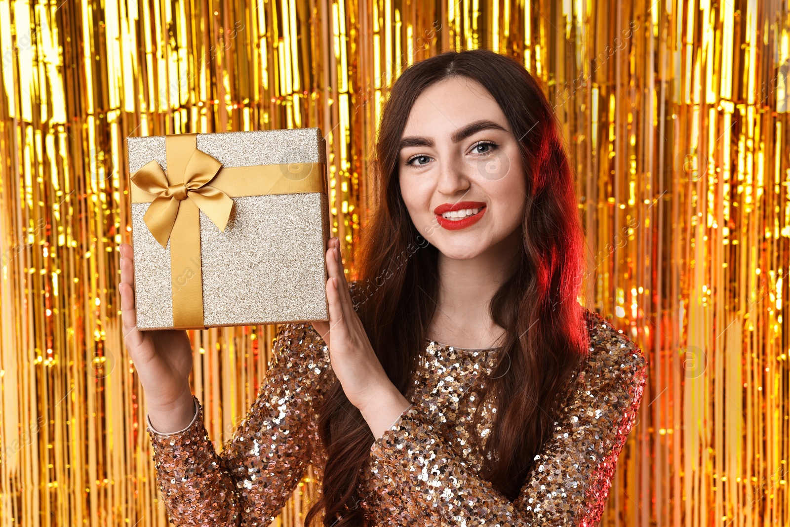 Photo of Happy young woman with gift box against golden foil curtain