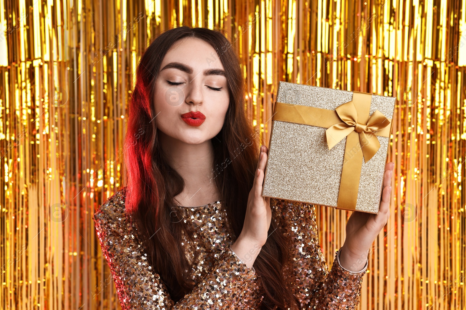 Photo of Beautiful young woman with gift box sending air kiss against golden foil curtain