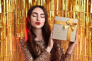 Photo of Beautiful young woman with gift box sending air kiss against golden foil curtain