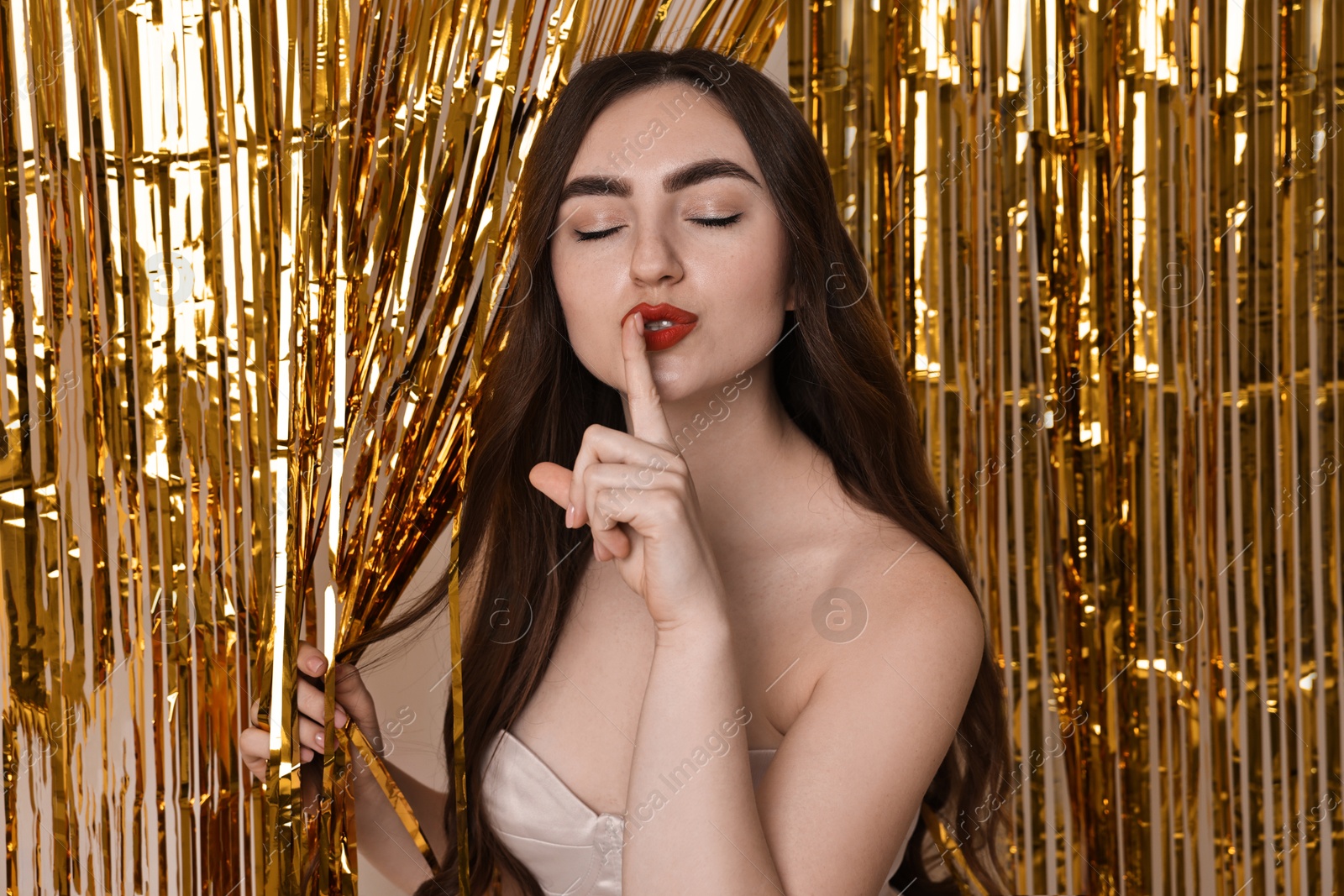 Photo of Beautiful young woman showing hush gesture near golden foil curtain against beige background