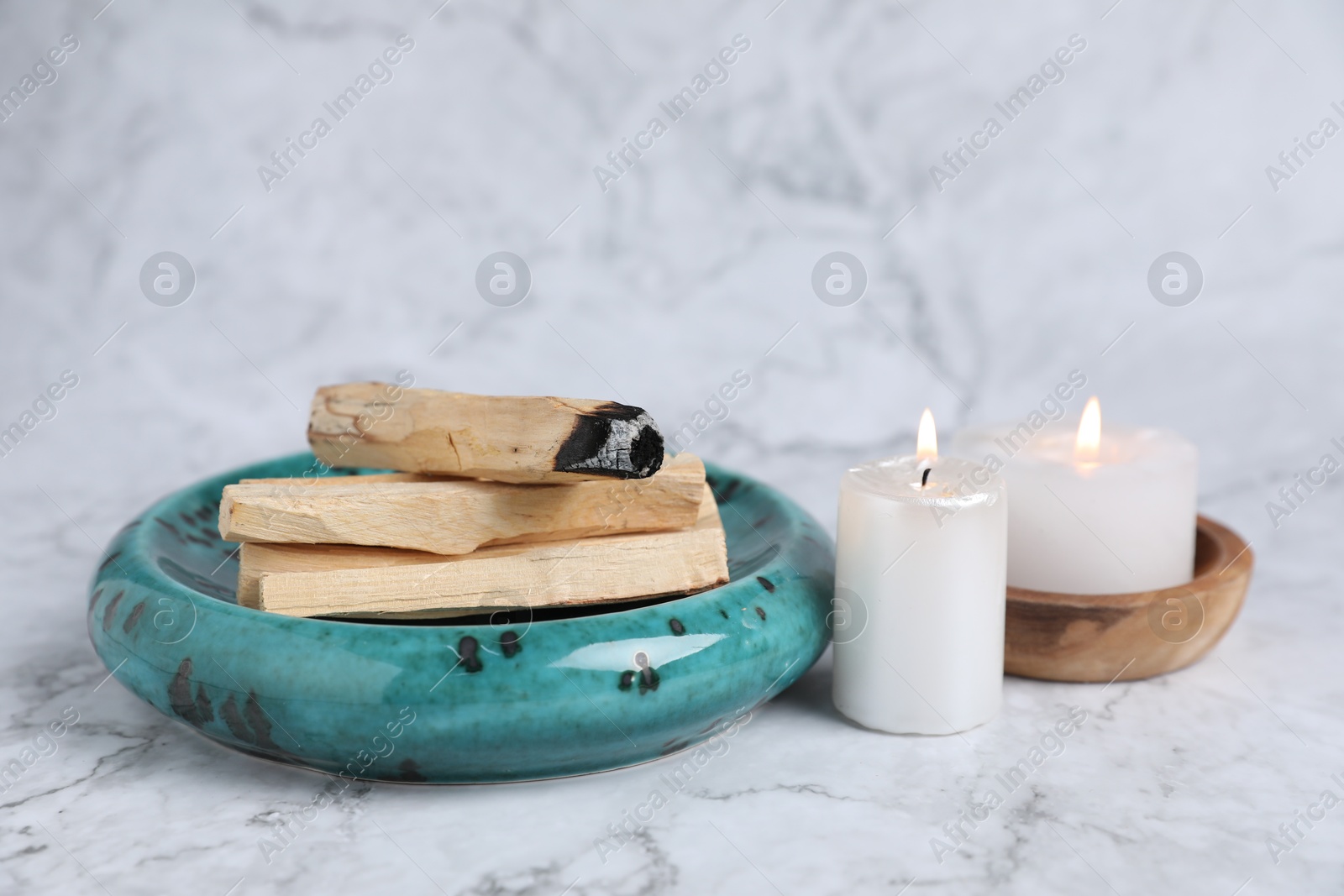 Photo of Palo santo sticks, burnt one and burning candles on white marble table, closeup