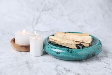 Photo of Palo santo sticks, burnt one and burning candles on white marble table, closeup