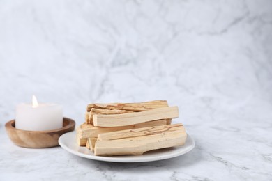 Photo of Palo santo sticks and burning candle on white marble table, closeup. Space for text