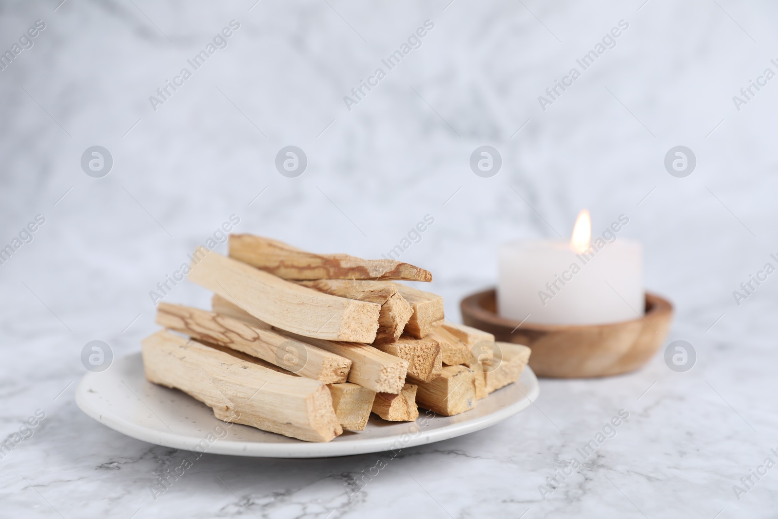 Photo of Palo santo sticks and burning candle on white marble table, closeup