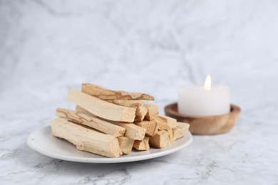 Photo of Palo santo sticks and burning candle on white marble table, closeup