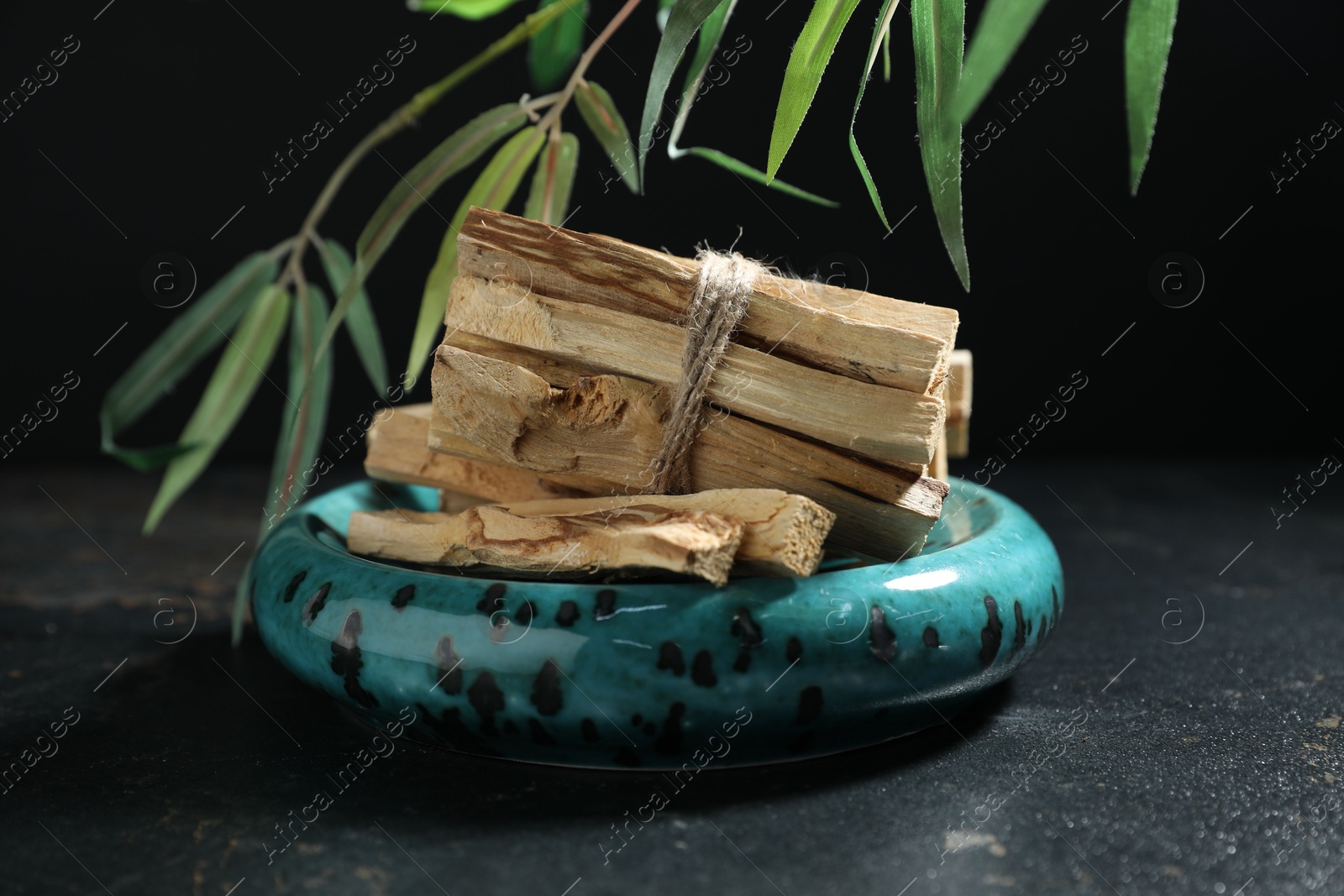 Photo of Bunch of palo santo sticks on dark table and green branch against black background, closeup