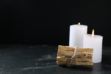 Photo of Bunch of palo santo sticks and burning candles on dark table, space for text