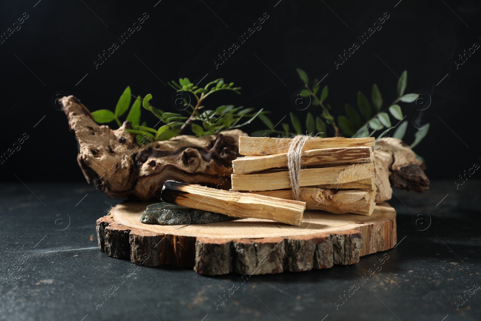 Photo of Smoldering palo santo stick, gemstone, snag and green branch on dark table, closeup