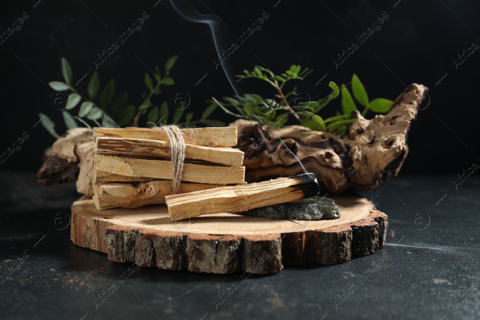 Photo of Smoldering palo santo stick, gemstone, snag and green branch on dark table, closeup