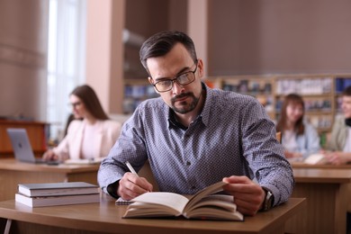 Handsome man reading book and taking notes at desk in public library