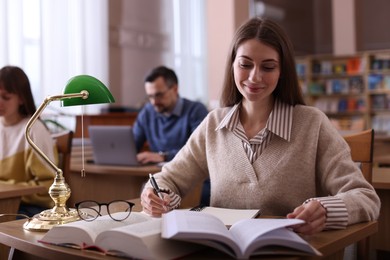 Beautiful woman reading book and taking notes at desk in public library