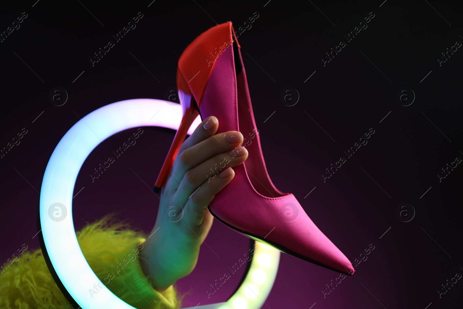 Photo of Woman holding high heeled shoe through ring lamp on dark purple background, closeup. Space for text