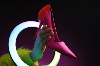 Photo of Woman holding high heeled shoe through ring lamp on dark purple background, closeup. Space for text