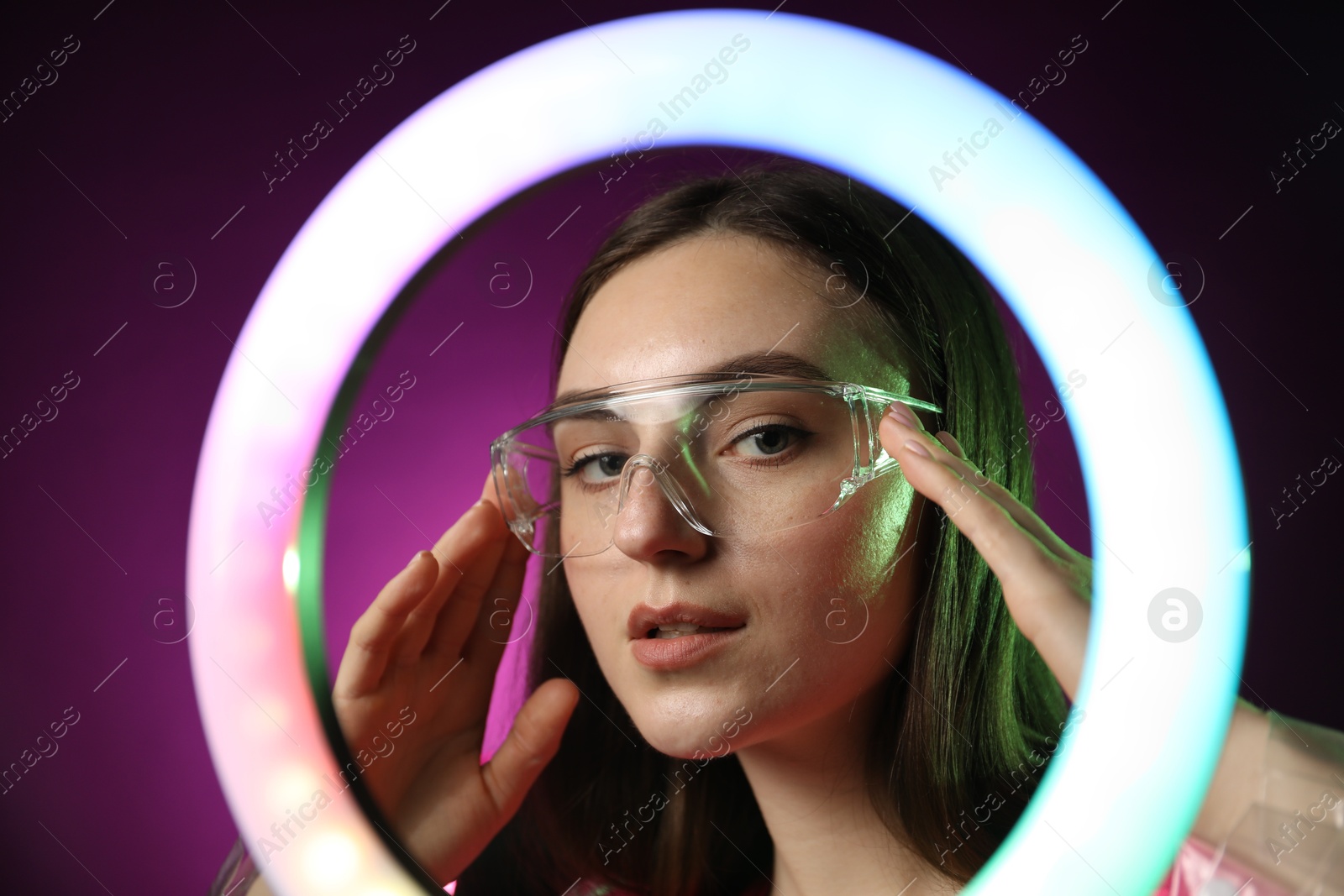 Photo of Stylish woman wearing glasses on dark purple background, view through ring lamp