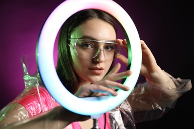 Photo of Stylish woman wearing transparent coat and glasses on dark purple background, view through ring lamp