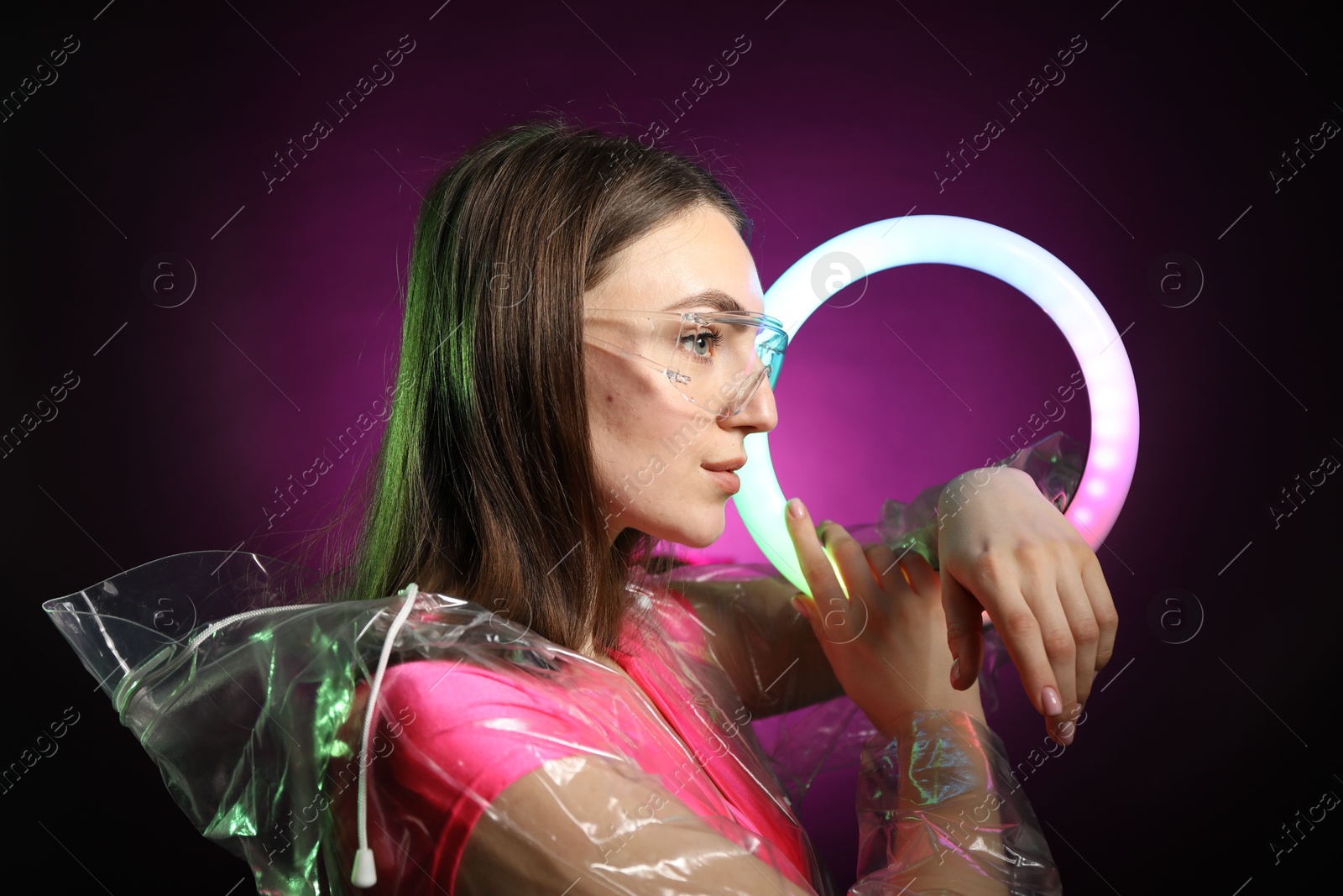 Photo of Stylish woman with ring lamp wearing transparent coat and glasses on dark purple background