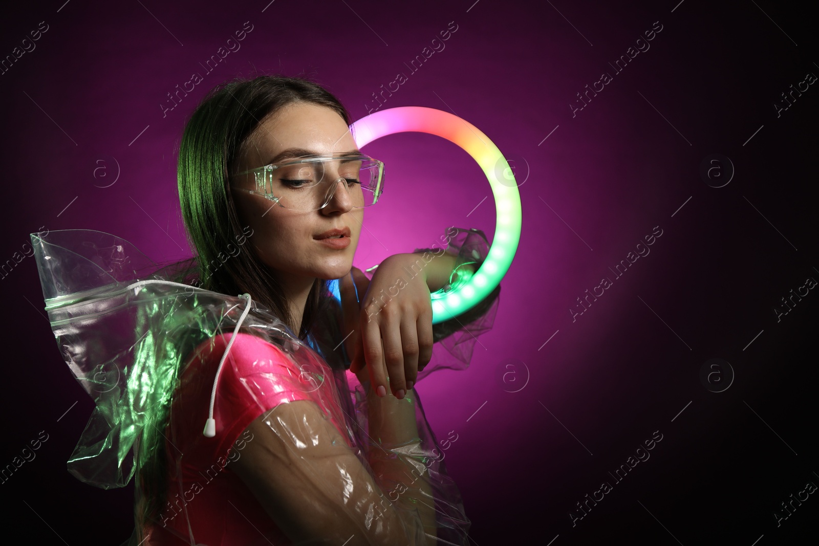 Photo of Stylish woman with ring lamp wearing transparent coat and glasses on dark purple background, space for text