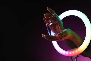 Photo of Woman holding perfume through ring lamp on dark purple background in neon lights, closeup. Space for text