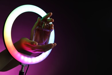 Photo of Woman holding perfume through ring lamp on dark purple background in neon lights, closeup. Space for text