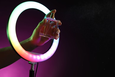 Photo of Woman holding perfume through ring lamp on dark purple background in neon lights, closeup. Space for text