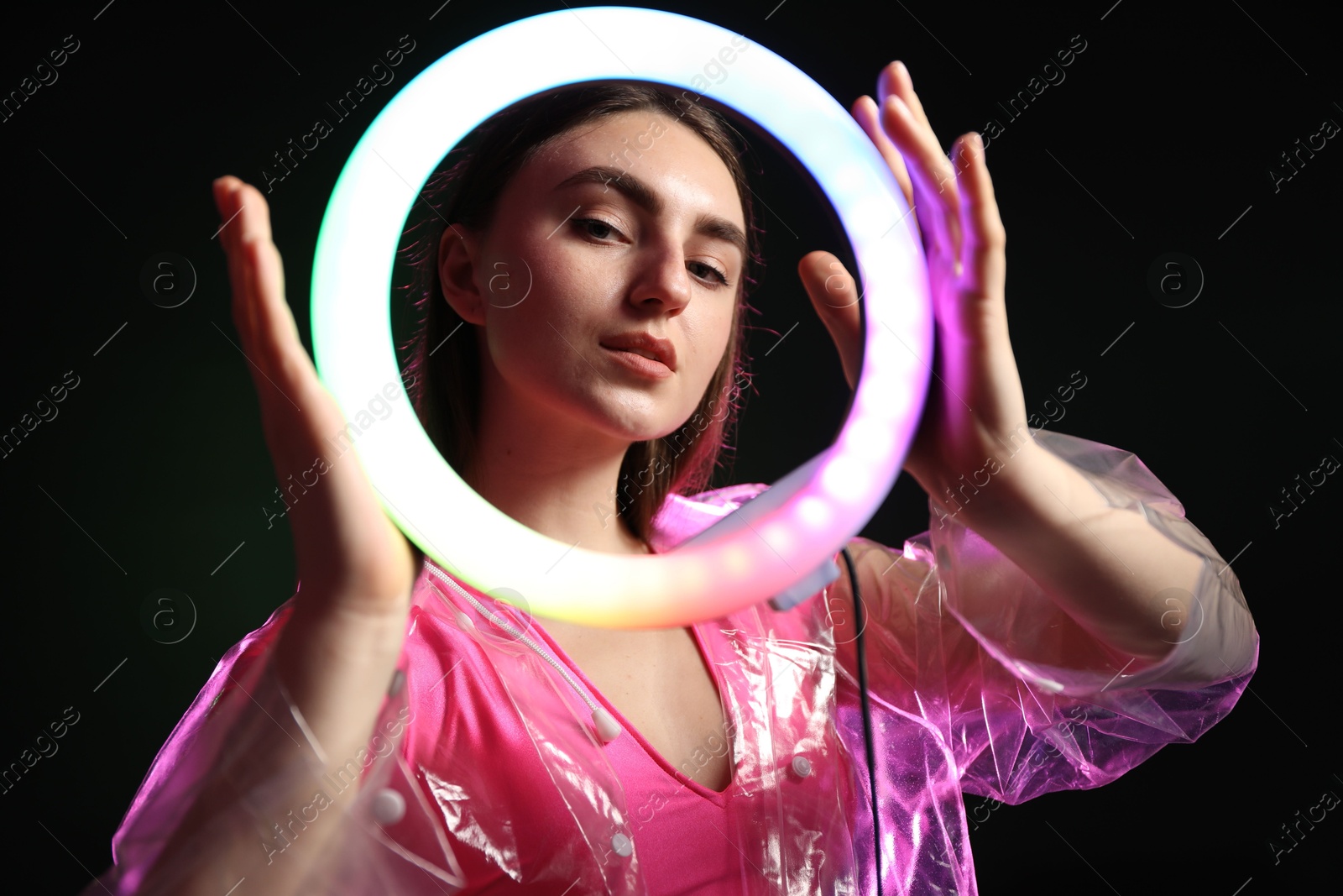 Photo of Stylish woman wearing transparent coat on dark purple background, view through ring lamp