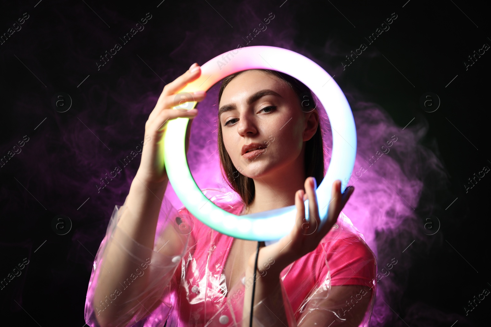 Photo of Stylish woman wearing transparent coat on dark purple background in smoke, view through ring lamp