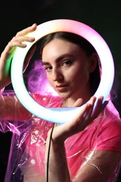 Photo of Stylish woman wearing transparent coat on dark purple background in smoke, view through ring lamp