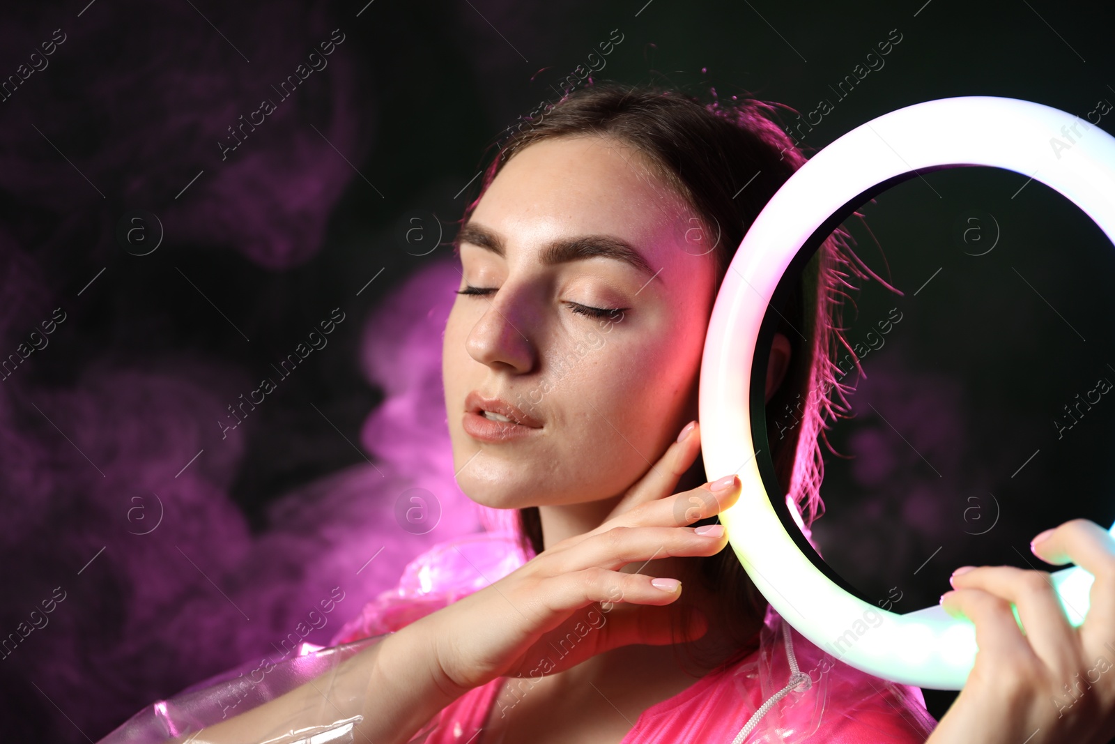 Photo of Beautiful woman with ring lamp wearing transparent coat on dark purple background in smoke, space for text