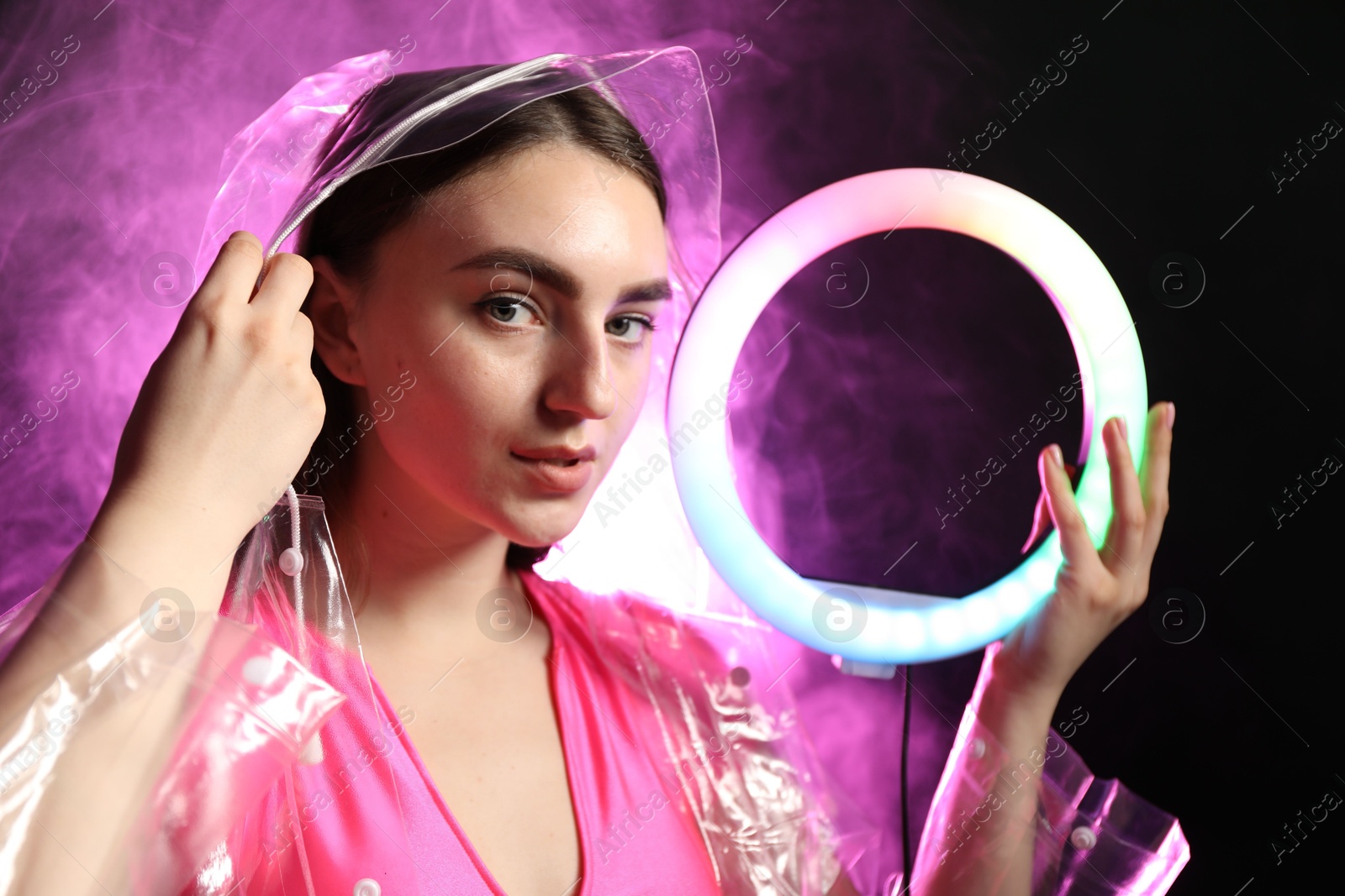 Photo of Beautiful woman with ring lamp wearing transparent coat on dark purple background in smoke