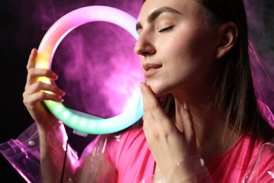 Photo of Beautiful woman with ring lamp wearing transparent coat on dark purple background in smoke