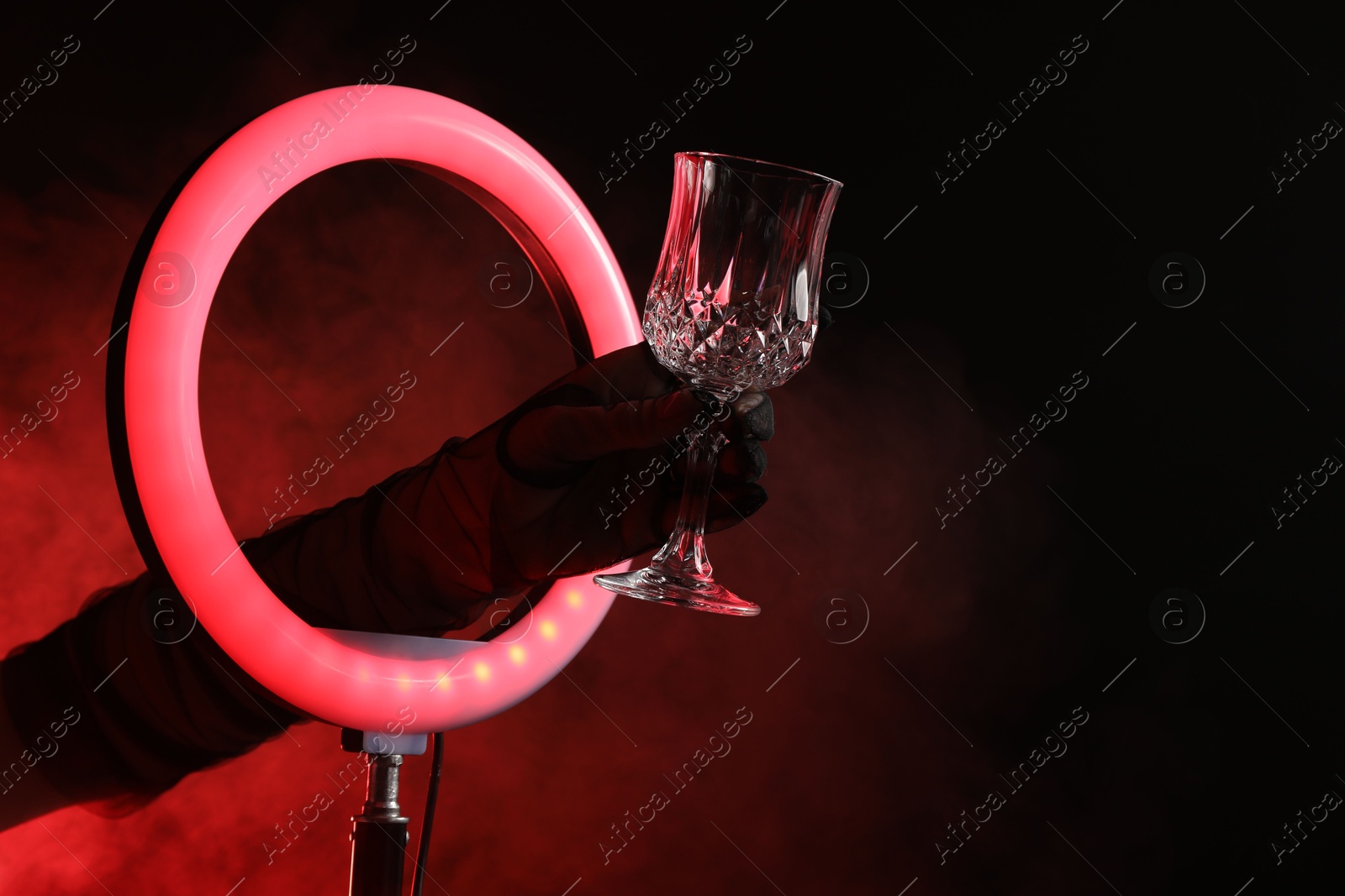 Photo of Woman holding empty wineglass through ring lamp on dark red background in smoke, closeup. Space for text