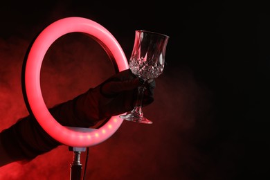 Photo of Woman holding empty wineglass through ring lamp on dark red background in smoke, closeup. Space for text