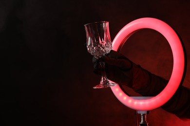 Photo of Woman holding empty wineglass through ring lamp on dark red background in smoke, closeup. Space for text