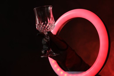 Photo of Woman holding empty wineglass through ring lamp on dark red background in smoke, closeup. Space for text