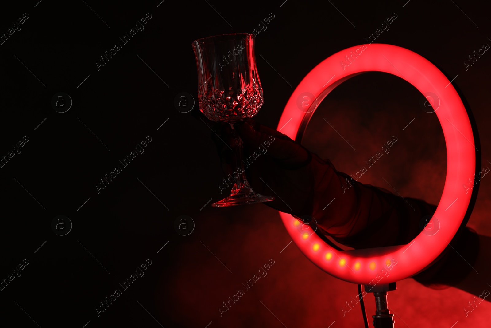 Photo of Woman holding empty wineglass through ring lamp on dark red background in smoke, closeup. Space for text