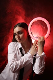 Photo of Beautiful woman with ring lamp on dark red background in smoke