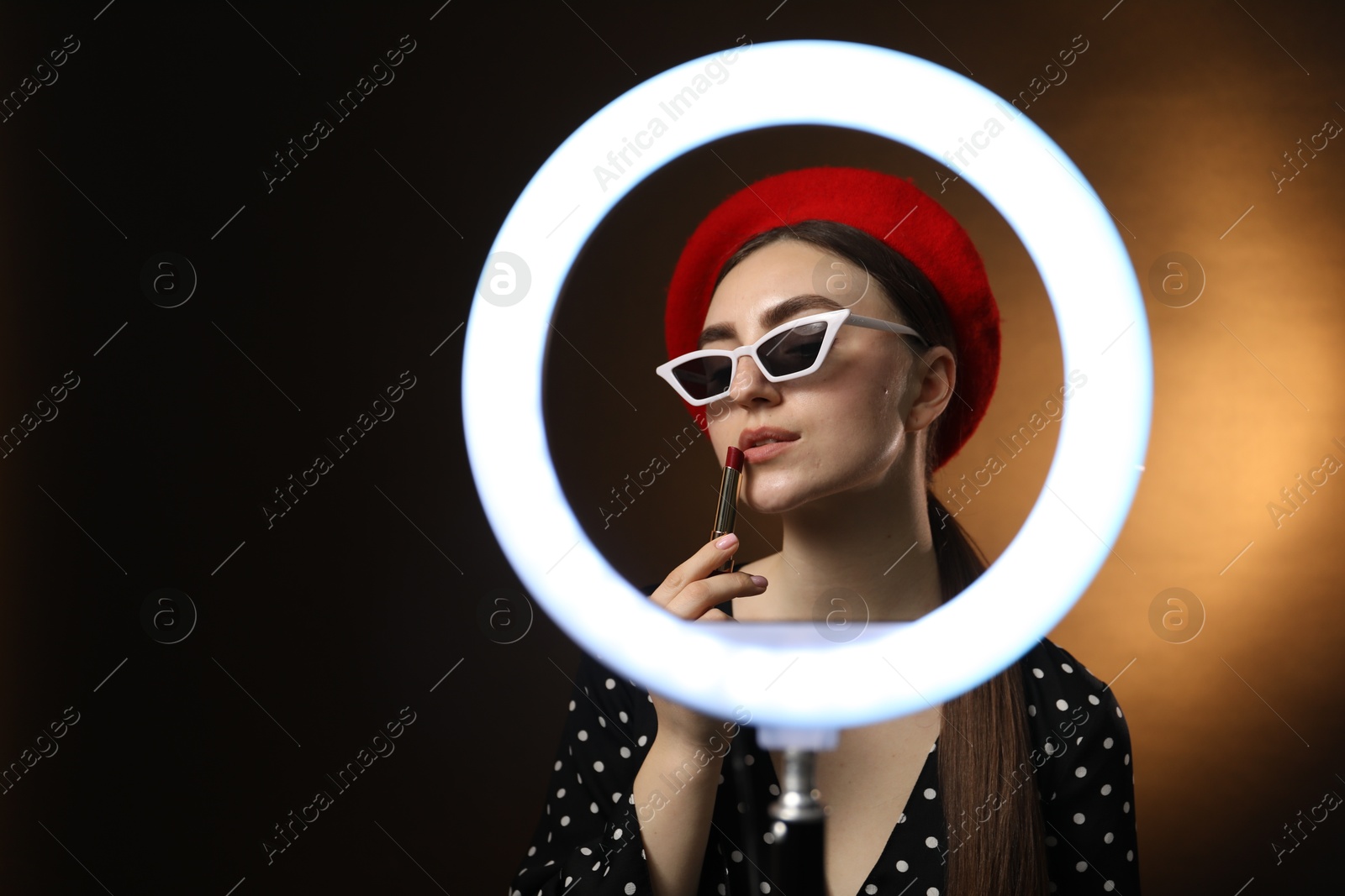 Photo of Beautiful woman applying lipstick on dark background, view through ring lamp. Space for text