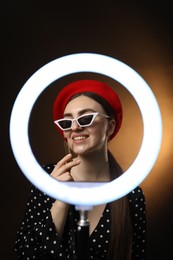 Photo of Beautiful woman applying lipstick on dark background, view through ring lamp