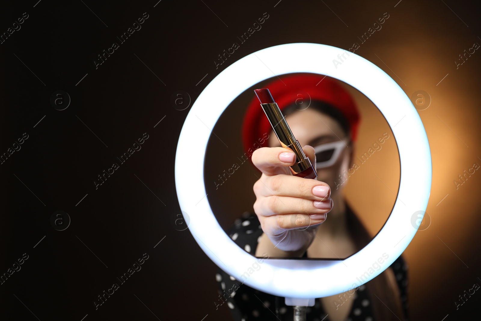 Photo of Beautiful woman holding lipstick through ring lamp on dark background, selective focus. Space for text