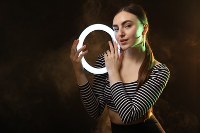 Photo of Beautiful woman with ring lamp on dark background in smoke, space for text