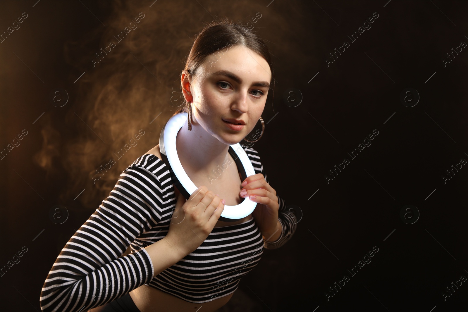 Photo of Beautiful woman with ring lamp on dark background in smoke, space for text