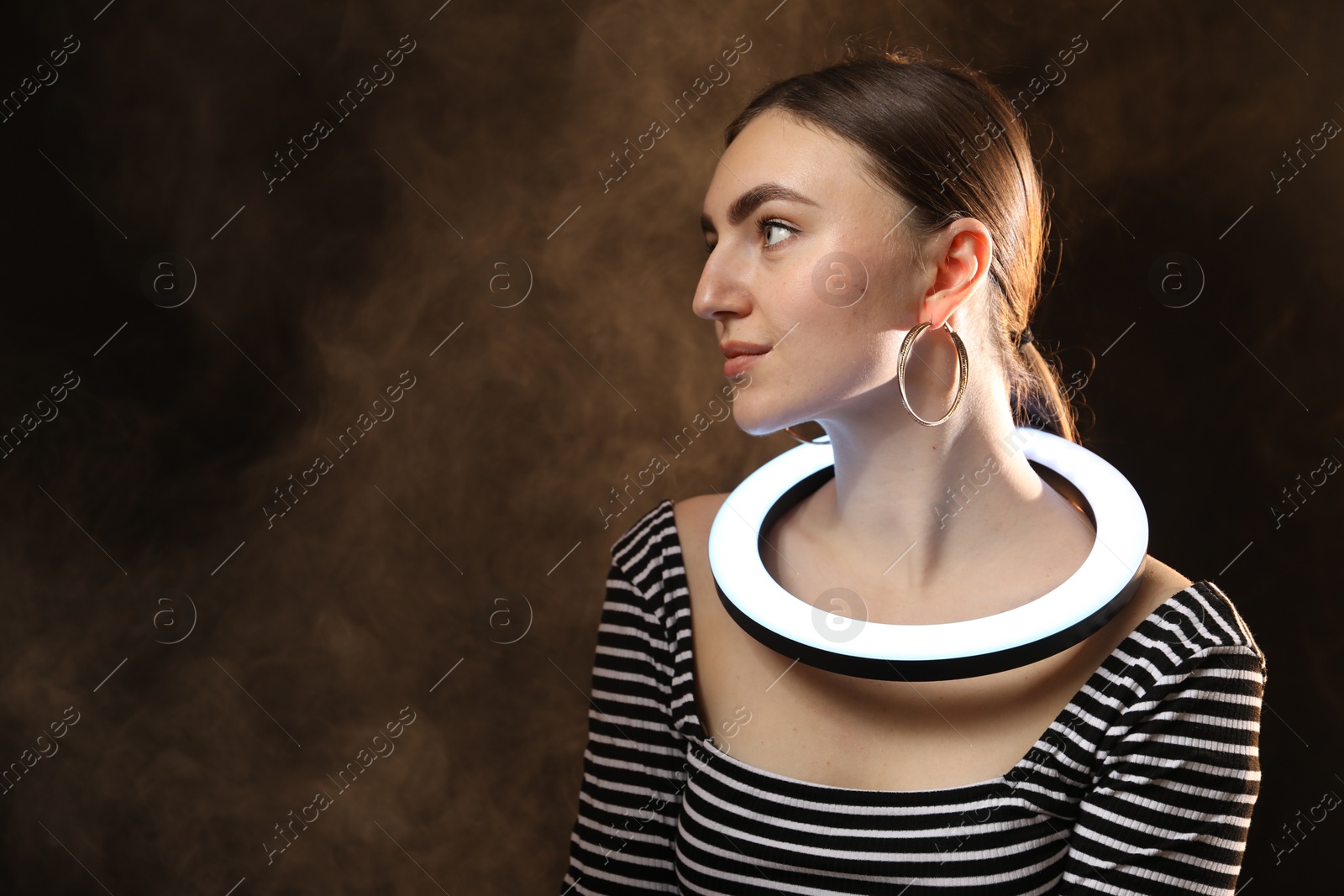 Photo of Beautiful woman with ring lamp on dark background in smoke, space for text