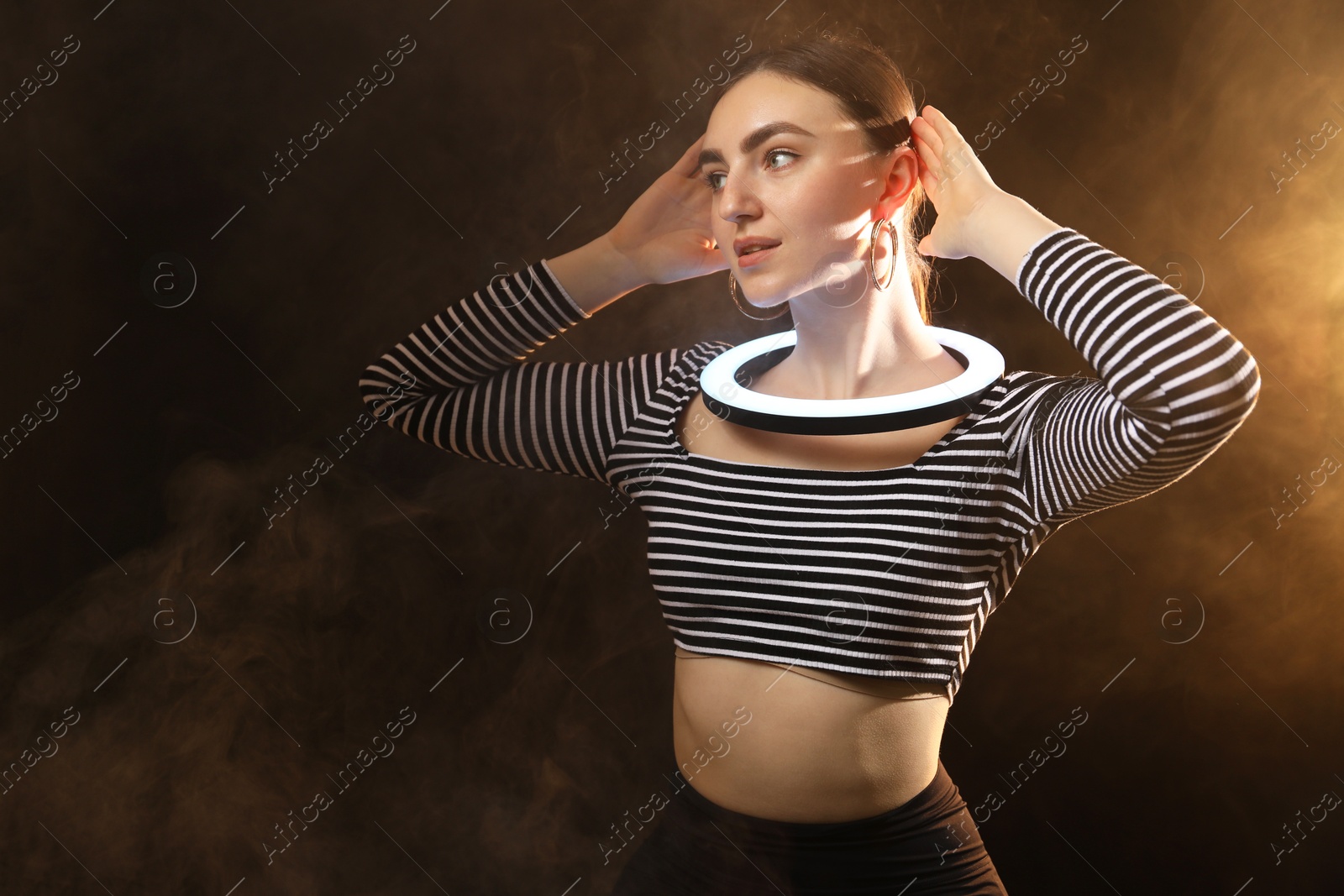 Photo of Beautiful woman with ring lamp on dark background in smoke, space for text